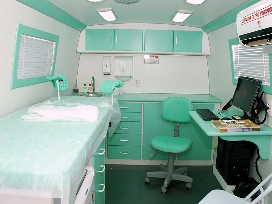 Interior de unidade móvel de saúde com maca de exame, bancada com pia, armários e mesa de atendimento